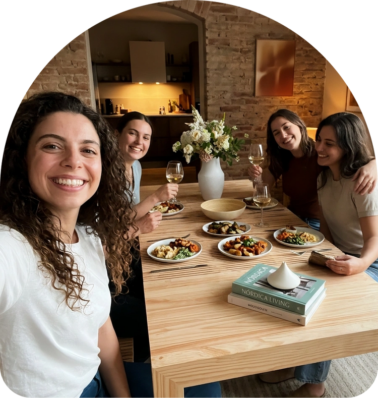 Four women smiling and posing together around a wooden dining table set with plates of food, wine glasses, and a vase of flowers in a cozy home kitchen setting.