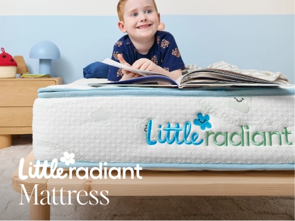 A child lying on a mattress in a bedroom setting, reading a book. The mattress has a white quilted top with a light blue border and the brand name 'Little radiant' printed on the front.
